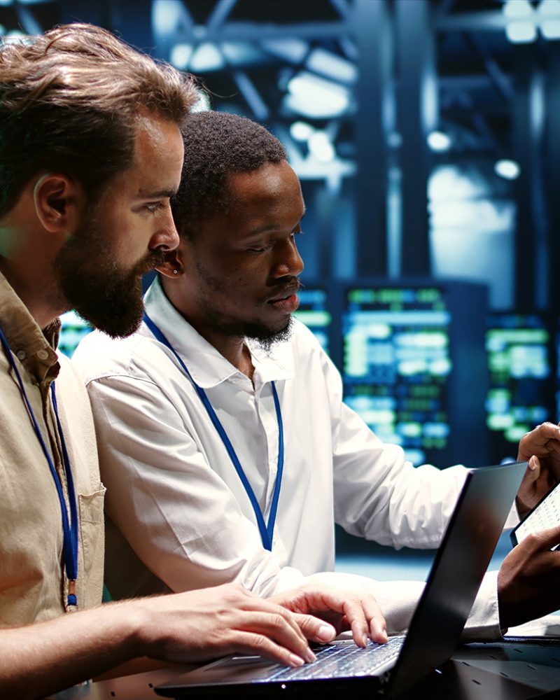 Coworkers running data center hardware diagnostic tests to determine and service software issues. Servicemen using laptop and tablet to fix critical systems, checking for blips
